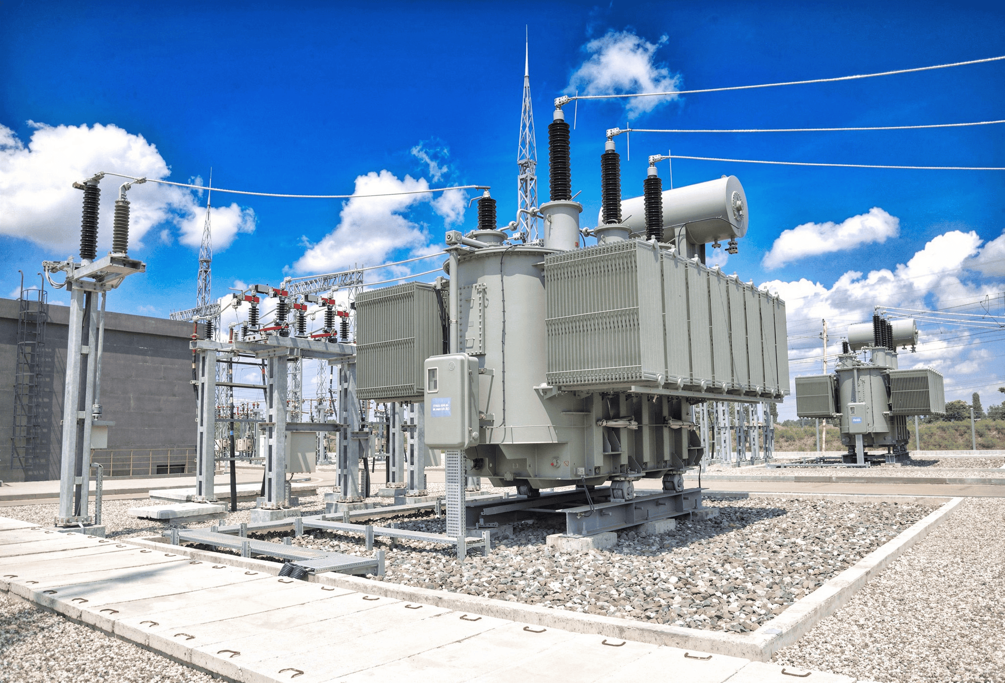 Large electrical transformers at a power substation under a clear blue sky with white clouds.